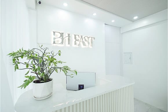 A minimalistic reception area with a curved white desk, a potted plant, and a sign reading "BH EAST" on the wall.