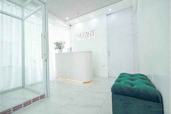 Minimalist white reception area with a sleek counter, a small potted plant, and a teal cushioned bench. The word "BH EAST" is illuminated on the wall above the counter.