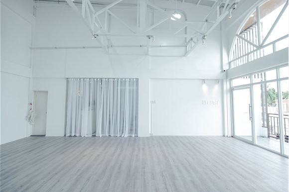 Spacious, empty room with white walls, high ceiling, and large windows. Light wood flooring and sheer white curtains on one side.