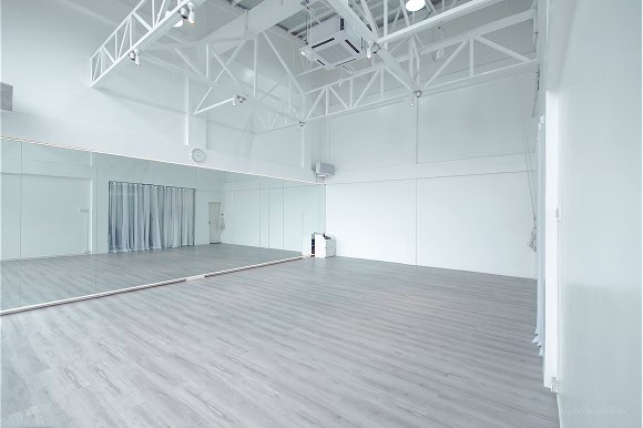 Empty dance studio with large wall mirrors, white walls, and a gray wooden floor. Ceiling has exposed beams and lights.