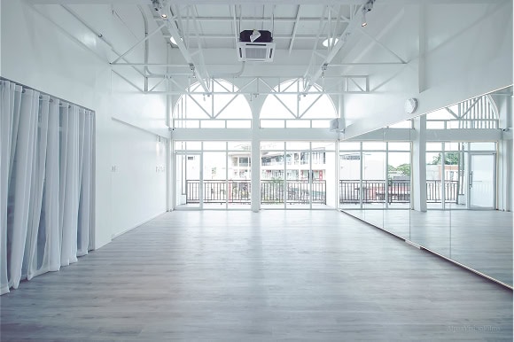 Spacious, empty dance studio with large windows, white walls, and wooden flooring. Ceiling has exposed beams and air conditioning unit.