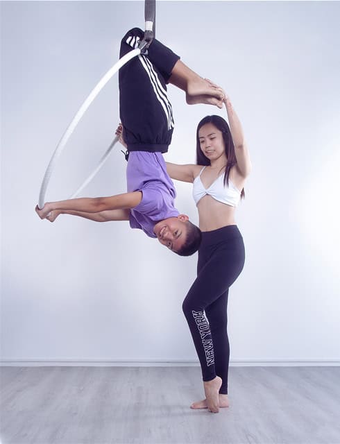 A woman assists a boy performing an aerial hoop pose. The boy is upside down, holding the hoop with his legs. The background is plain white.