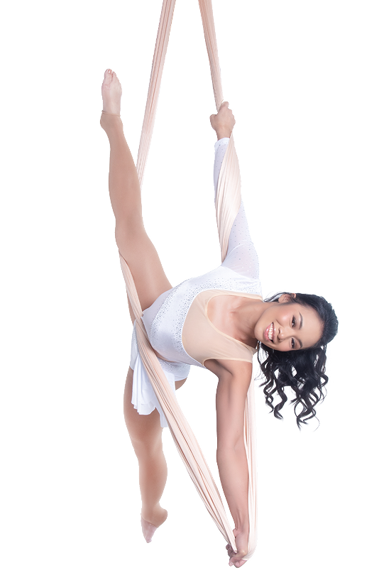 A woman in a white leotard performs an aerial silk pose, holding the fabric with both hands while extending one leg upward.